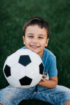 portrait of happy boy sitting on football field. sports section. Training of children, child with football soccer ball on field. soccer champion. Sport concept. kid laying downの写真素材
