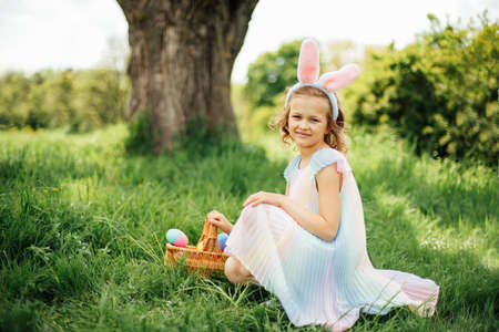 Easter egg hunt. Girl child Wearing Bunny Ears Running To Pick Up Egg In Garden. Easter tradition. Baby with basket full of colorful eggs. Bunny in basketの写真素材