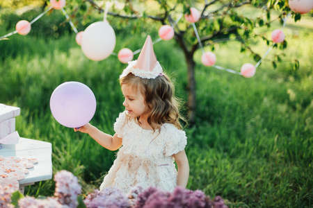 Happy birthday little girl making wish blowing candles on cake with pink decor in beautiful garden. 4 years birthday. child eat happy birthday pink cupcake. colorful pastel decoration outdoorの写真素材