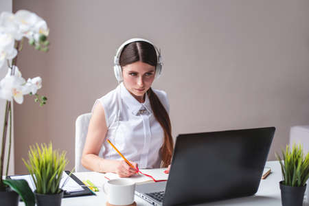 Focused millennial woman listening educational webinar, taking notes, getting remote online education at home. Female freelancer working on remote house. Woman in headphones learning languageの写真素材