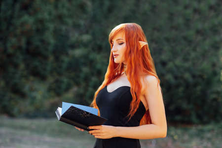 Close up portrait of Halloween Beautiful young Witch woman on spooky dark magic forest background with magic Book of spells. redhead girl with big eyes magical sightの写真素材
