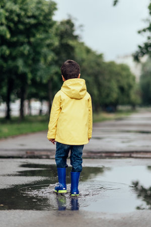 Little boy in raincoat and rubber boots playing in puddle. Fun on street. body hardening in summer. Splashes, drops of water, outdoor. waterproof boots jump in puddle and mud in the rain.の写真素材