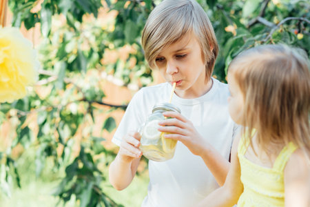 Children drink natural lemonade at stand in park. Summer refreshing natural drink lemonade. Detox fruit infused flavored water, cocktail in a beverage dispenser with fresh fruitsの写真素材