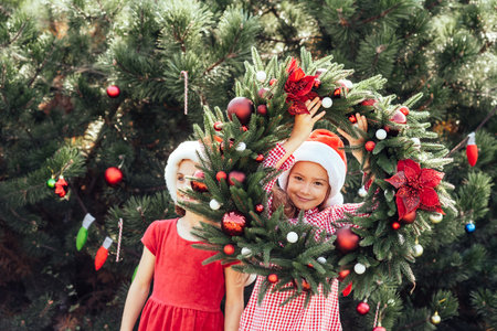 Merry Christmas. Portrait of two happy funny children girls in Santa hat with Christmas wreath. Happy Holidays. Fairy Magic. Happy kids enjoying holiday. Christmas in Julyの写真素材