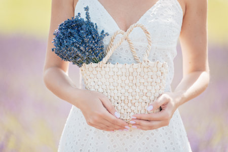 woman in white dress standing and holding straw bag with lavender flowers in her hand. Woman in Lavender Field at Sunsetの写真素材