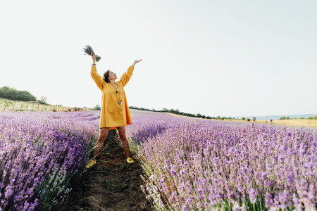 Woman in a yellow dress holding a lavender bouquet, spreading her arms in a lavender field, capturing a moment of freedom and joy.の写真素材