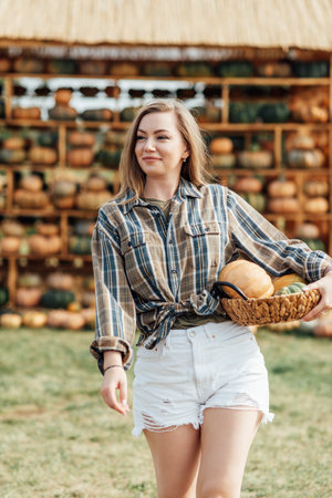 Young woman holding a basket of pumpkins near a haystack on a farm field. Outdoor autumn harvest scene. Fall season and agriculture concept for design and printの写真素材