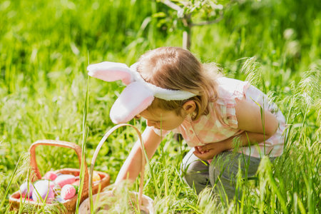 Easter egg hunt. Girl child Wearing Bunny Ears Running To Pick Up Egg In Garden. Easter tradition. Baby with basket full of colorful eggs.の写真素材