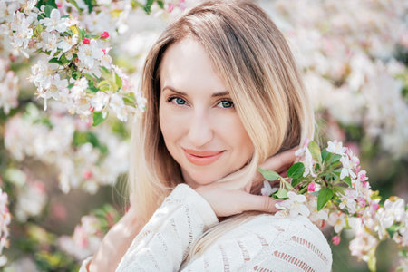 Portrait joyful young woman with blonde hair gently holds a cluster of white blossoms near her face...