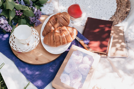 Cozy Outdoor Picnic with Croissants and Lilac Flowers charming picnic setup with croissants, tea, lilac flowers, books, and pastries on a blanket under dappled sunlight in spring dayの写真素材