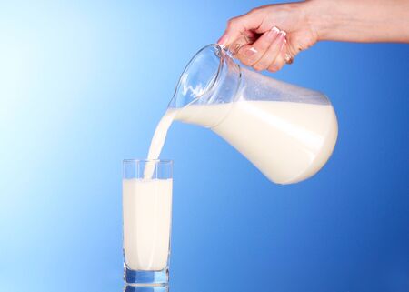 Someone pours milk from a jug into a glass on blue backgroundの写真素材