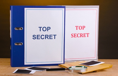 Envelopes and folders with top secret stamp and magnifying glass with photo papers on wooden table on brown backgroundの写真素材