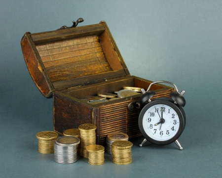 Alarm clock with coins in chest on grey background