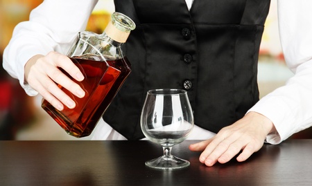 Barman hand with bottle of cognac  pouring drink into glass, on bright background