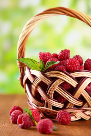 Ripe sweet raspberries in basket on wooden table, on green background