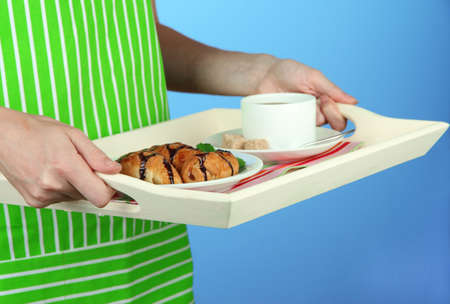 Woman in green apron holding wooden tray with breakfast, on color background