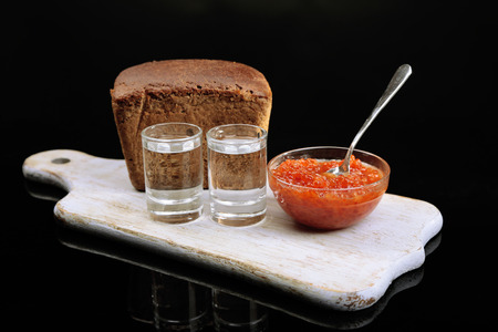 Composition with glass of vodka, red caviar, fresh bread on wooden board,  isolated on black
