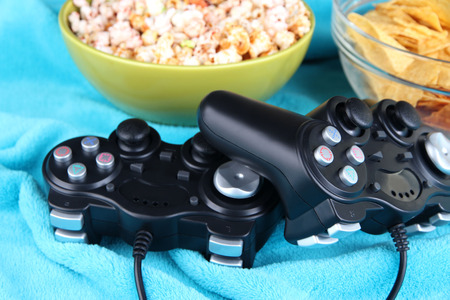 Black game controllers and bowl with snacks on color plaid background