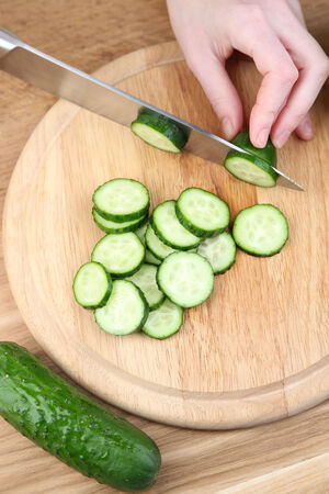 Female hands cutting cucumber on wooden board, close-up, on wooden backgroundの写真素材