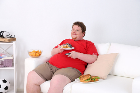 Lazy overweight male sitting with fast food on couch and watching televisionの写真素材