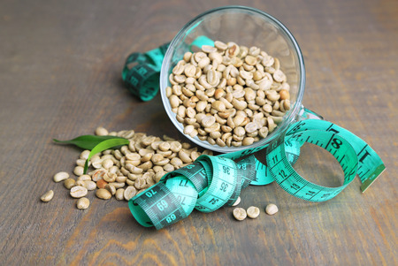 Raw green coffee beans in glass cup and measuring tape, on color wooden background. Concept of weight lossの写真素材