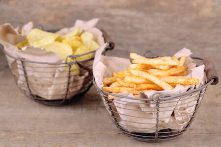 Tasty french fries in metal basket and potato chips on wooden table