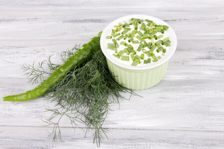 Round plastic bowl of cream with a tuft of dill and pepper near it on wooden backgroundの写真素材