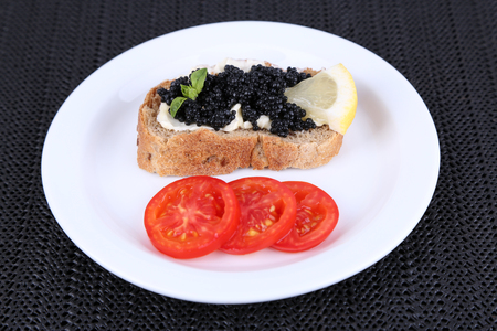 Black caviar and butter on bread with tomato on plate on dark fabric backgroundの写真素材