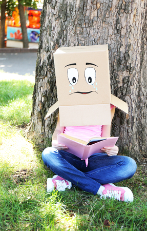 Woman with cardboard box on her head with sad face reading the book, outdoorsの写真素材