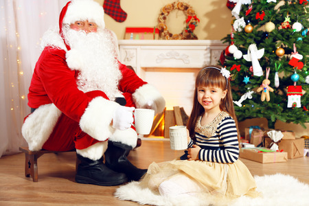 Santa Claus drinking hot chocolate with little cute girl near Christmas tree at homeの写真素材