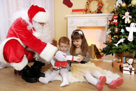 Santa Claus giving  present to  little cute girls near  fireplace and Christmas tree at homeの写真素材