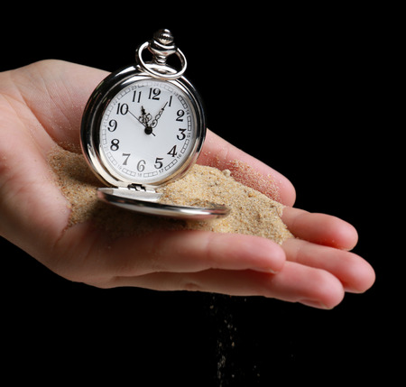 Silver pocket clock in hands and sand flowing away on black background