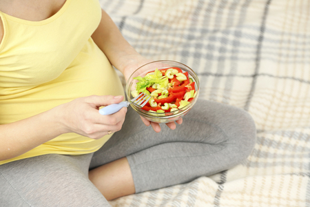 Young pregnant woman sitting on sofa with saladの写真素材