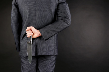 Businessman holding knife behind his back on gray backgroundの写真素材