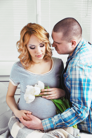Young pregnant woman sitting with husband and holding flowers in roomの写真素材