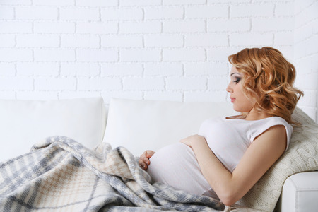 Young pregnant woman lying on sofa on light backgroundの写真素材