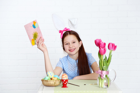 Cute girl with greeting card and decorated Easter eggs, on light backgroundの写真素材