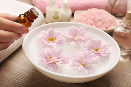 Female hand with bottle of essence and bowl of spa water with flowers on wooden table, closeupの写真素材