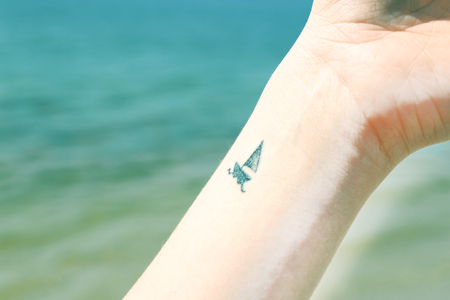 Close up of wrist of young woman with ship tattooed in it, on sea backgroundの写真素材