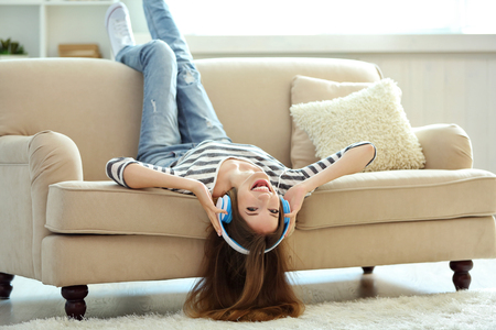 Woman listening music in headphones while lying on sofa in roomの写真素材