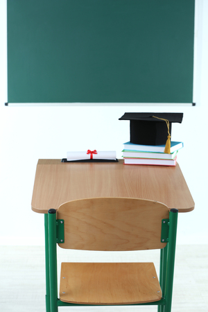 School desk with master hat, stack of books and diploma in classroomの写真素材