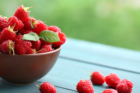 Fresh raspberries in bowl on wooden table on blurred nature background