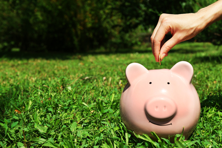 Female hand putting coin into pink piggy bank over green grass backgroundの写真素材