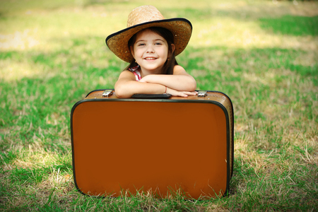 Happy little Caucasian girl in hat sitting behind suitcase, Outdoor portraitの写真素材