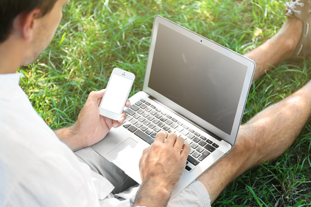 Man with laptop and cellphone sitting on green grass in the park, close upの写真素材
