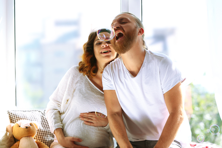 Happy beloved man and pregnant woman blow soap bubbles in waiting for baby's birth on window boardの写真素材