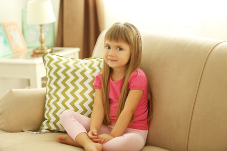 Beautiful little girl sitting on sofa, on home interior backgroundの写真素材
