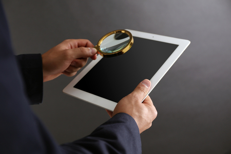 Businessman holding magnifying glass and digital tablet on dark grey background, searching and examining conceptの写真素材