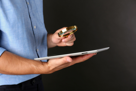Businessman holding magnifying glass and digital tablet on dark grey background, searching and examining conceptの写真素材