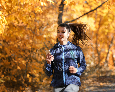Young beautiful woman running in autumn park and listening to music with headphones.の写真素材